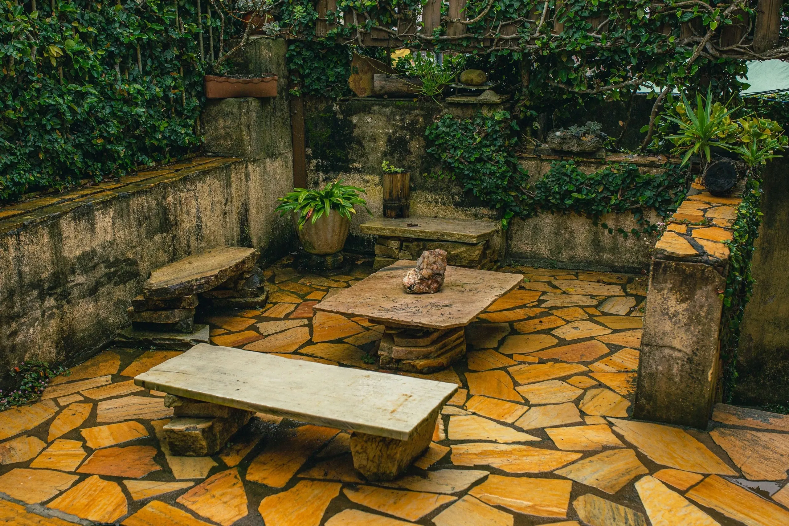 Une cour dallée de pierres irrégulières, avec un banc et une table en pierre, entre des murs couverts de lierre
